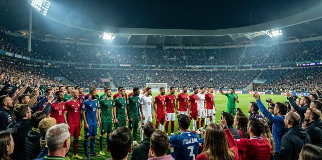 Stade de football européen rempli de supporters, avec des joueurs de sélections nationales alignés sur la pelouse sous les projecteurs, dans une ambiance nocturne réaliste.