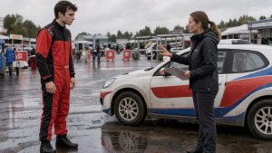 Jeune pilote en combinaison de course écoutant une instructrice près d’une voiture de rallye sur un circuit français.